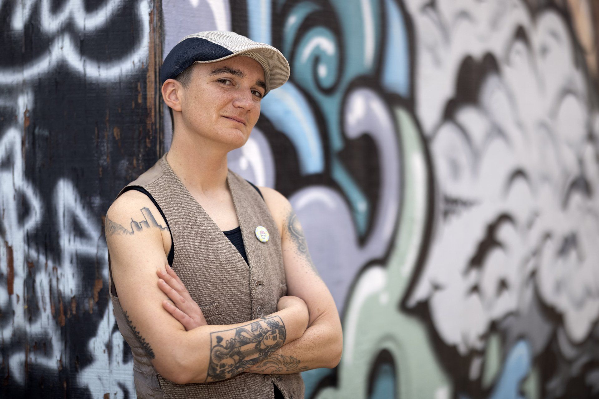 kim g standing against a graffitied wall, arms crossed, wearing a ball cap and a tweed vest.