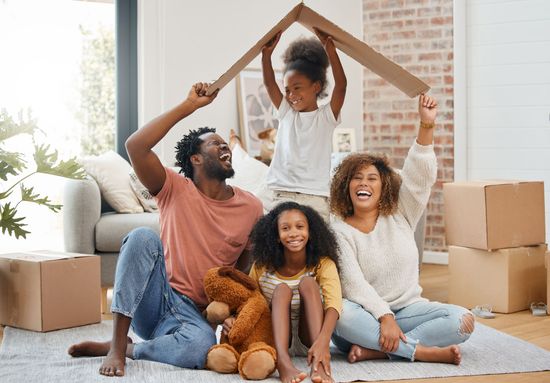 A family is sitting on the floor in a living room holding a cardboard house.