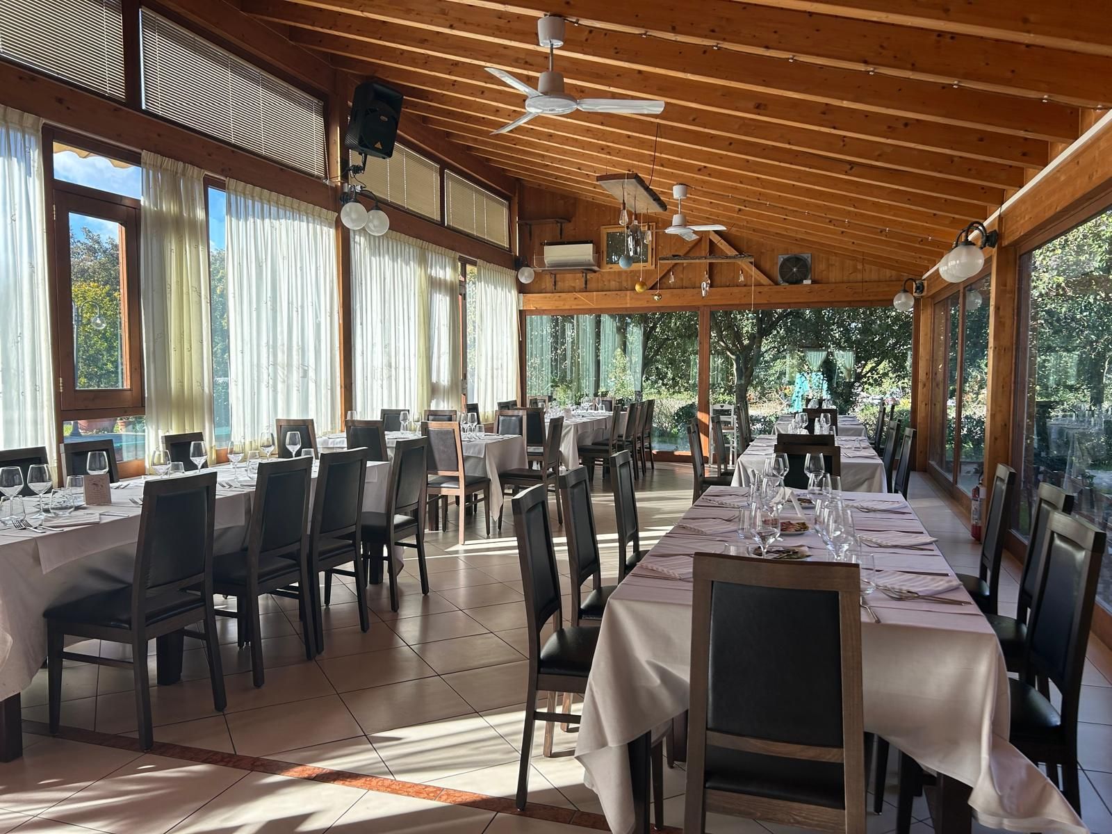 Sala da pranzo vuota di un ristorante con tavoli apparecchiati, sedie scure e grandi finestre sotto un soffitto di legno.
