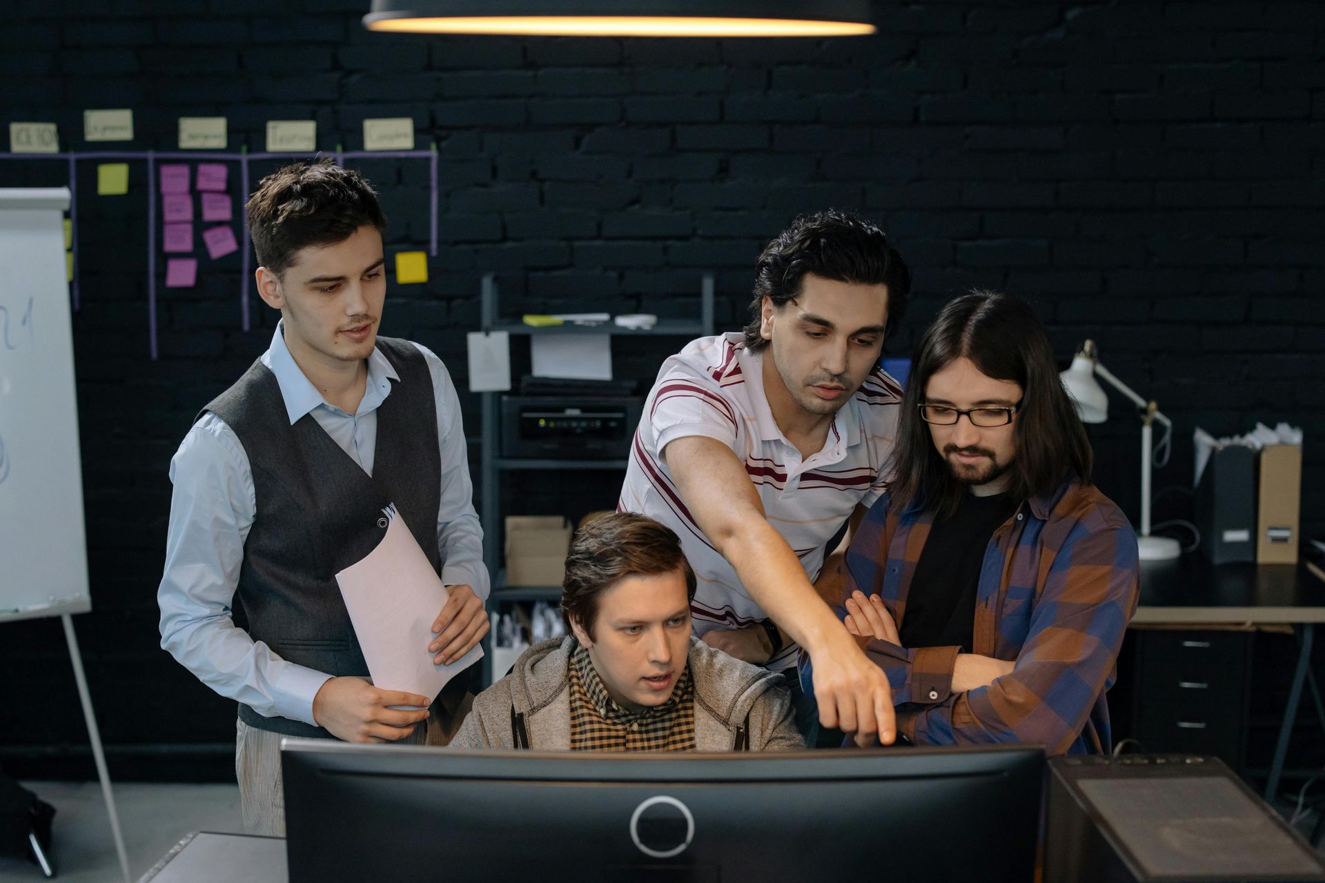 Four people gather around a computer monitor in an office, discussing and pointing.