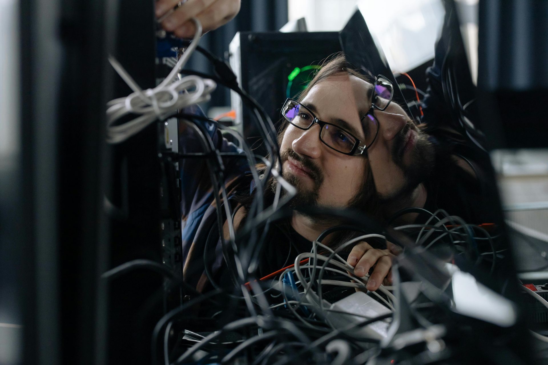 Man surrounded by wires, wearing glasses and headset, looking up with a thoughtful expression.