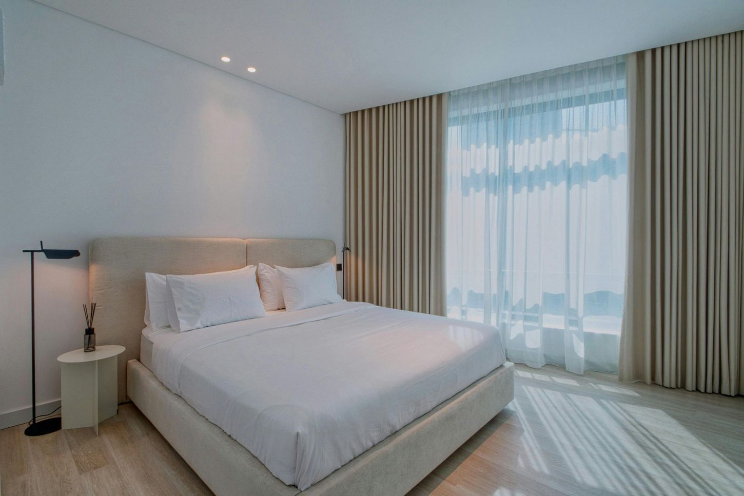 A modern, minimalist bedroom with a large bed, cream-colored curtains, and a sleek floor lamp on a light wood floor.