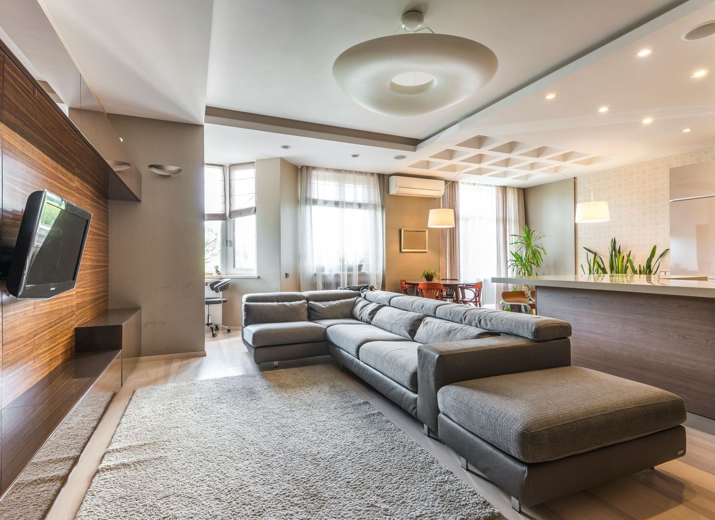 A modern living room with a large gray sectional sofa on a patterned rug, facing a mounted TV and open kitchen area.