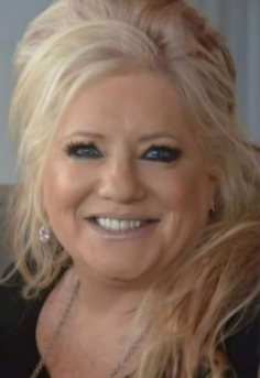 Blonde woman with blue eyes smiles, wearing black, in a soft-lit indoor setting.