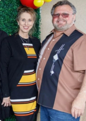 Woman and man pose together; she wears a patterned dress and a black cardigan; he wears a brown shirt with black panels.