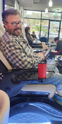 Man in a nail salon, getting a pedicure. He's holding a tablet, with a drink on a tray.