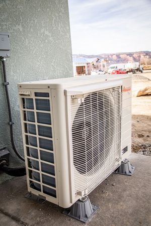Exterior air conditioning unit, light beige, against a stucco wall, mounted on gray supports.