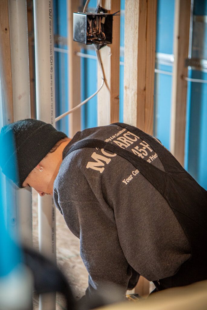 Person in a dark cap and gray hoodie works on electrical wiring in a framed wall with PVC pipes.