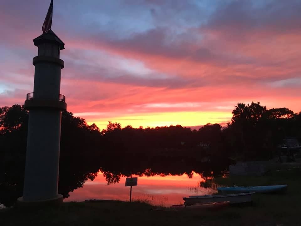 Lighthouse silhouetted against a vibrant sunset reflected in a calm lake.