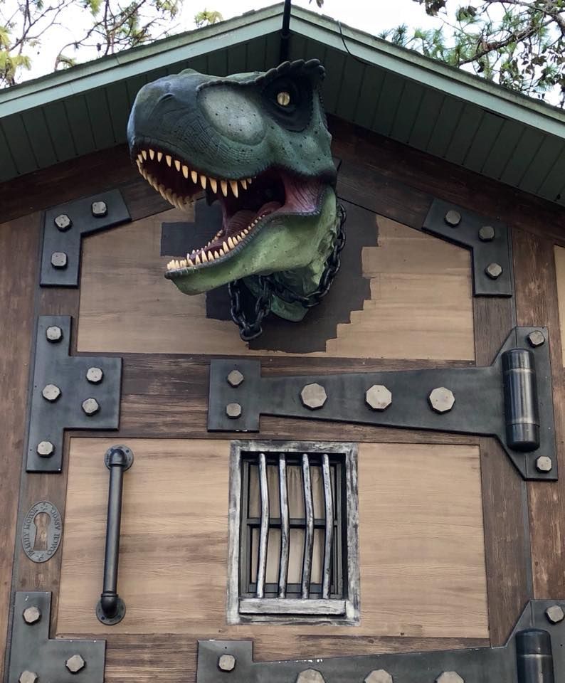 Dinosaur head mounted on a wooden building with metal details and a barred window.