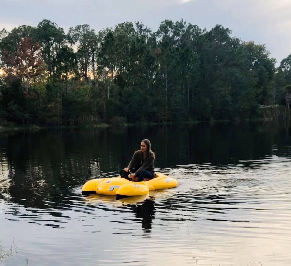 Woman on a yellow watercraft in a dark lake, surrounded by trees.