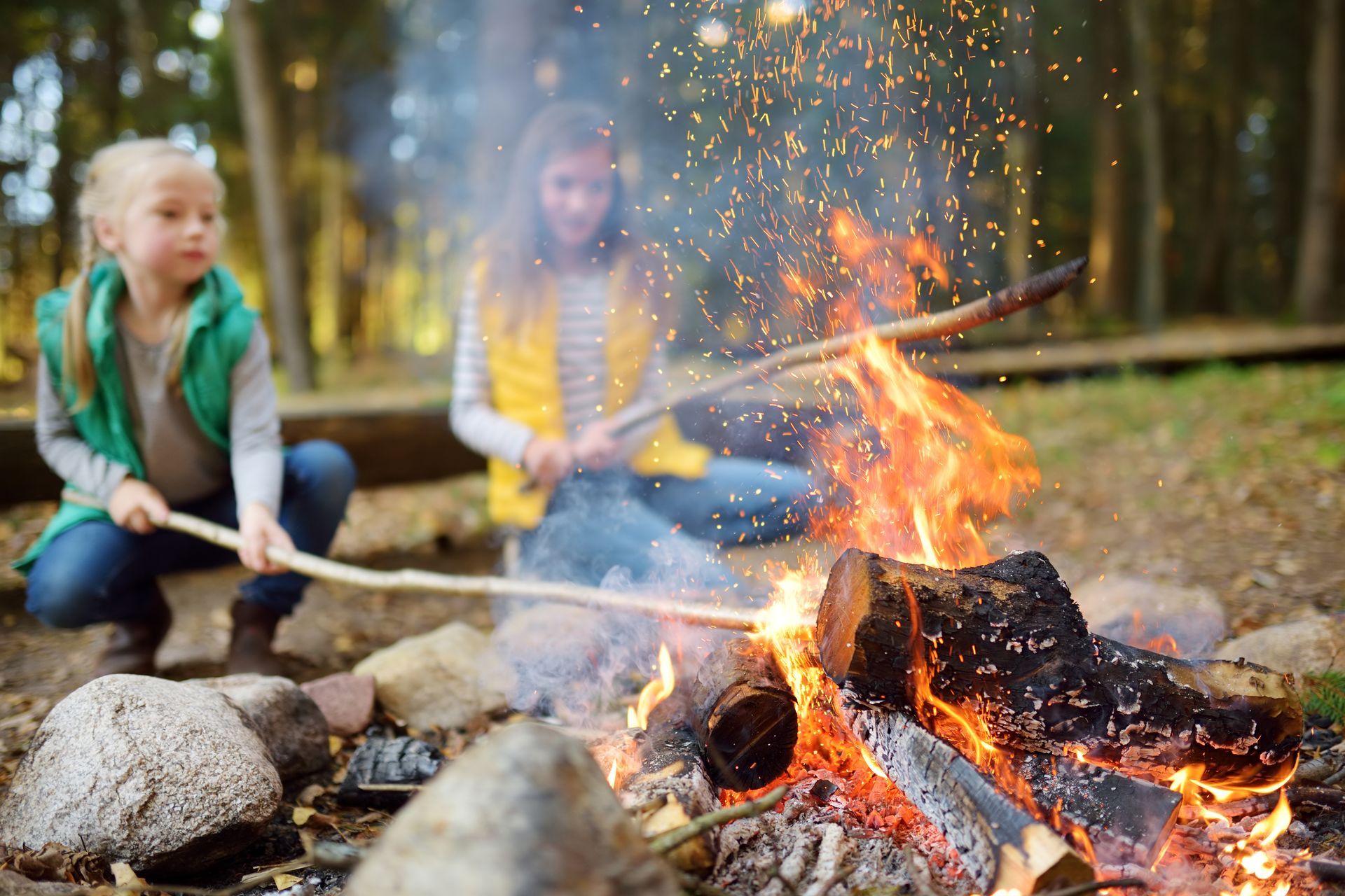 Two people sit by a campfire, holding sticks. Sparks fly.