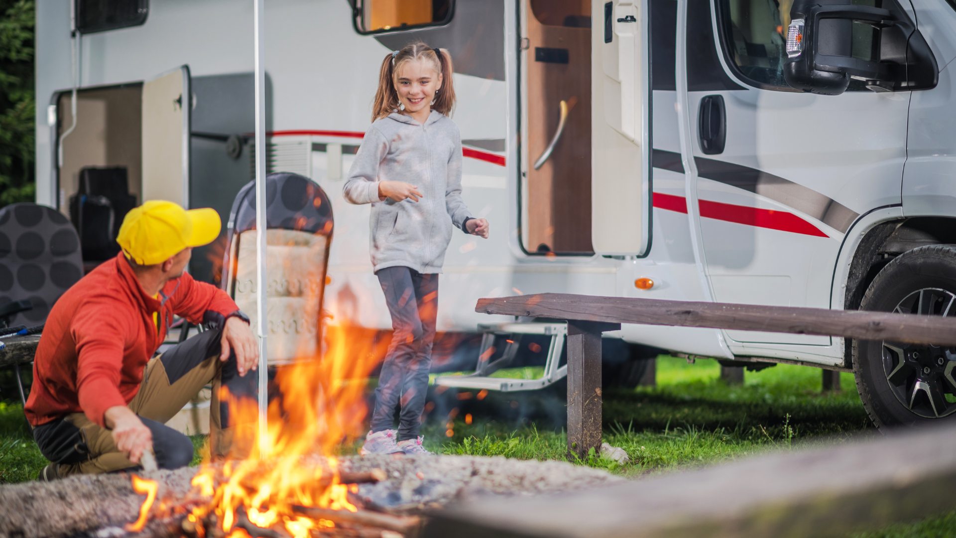 Man tending a campfire while girl dances near a white RV outdoors on a sunny day.
