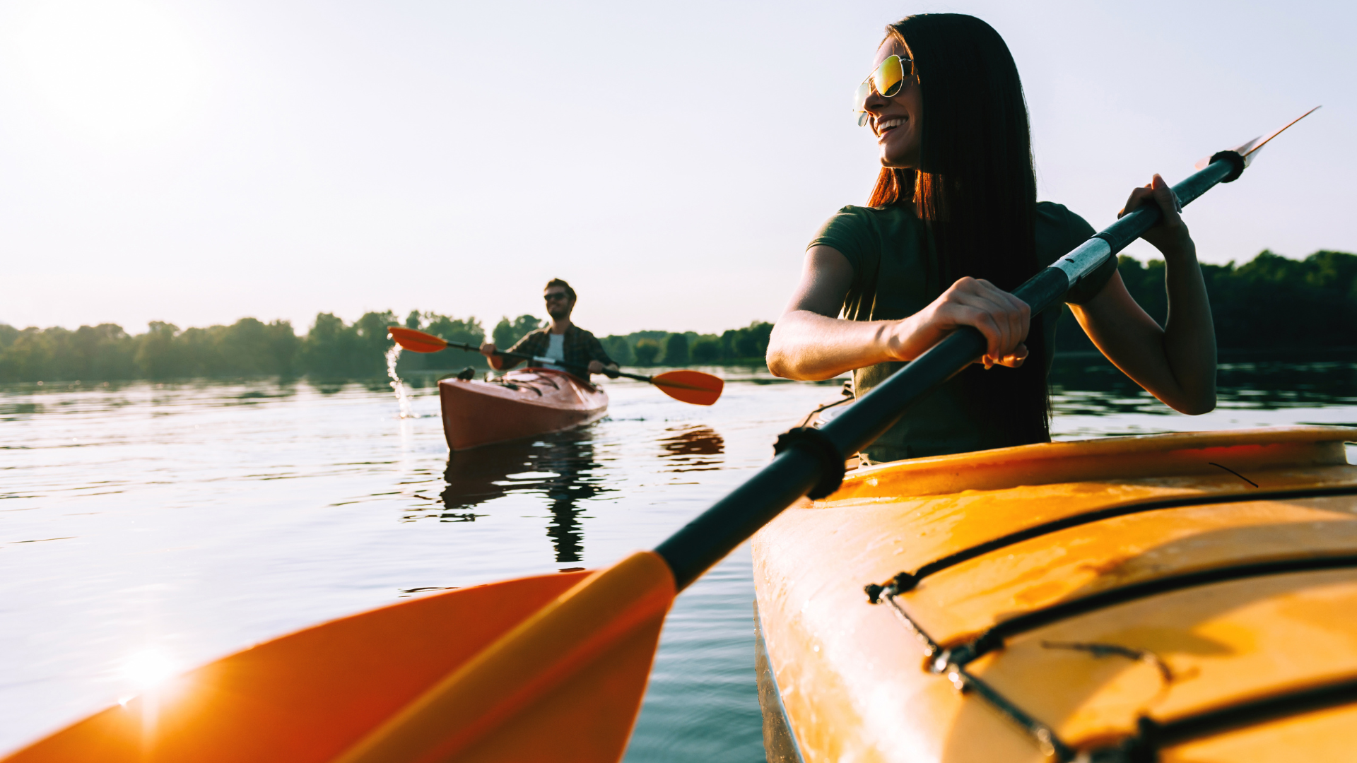 Woman kayaking on lake, holding paddle. Another person kayaks in background. Sun shines.