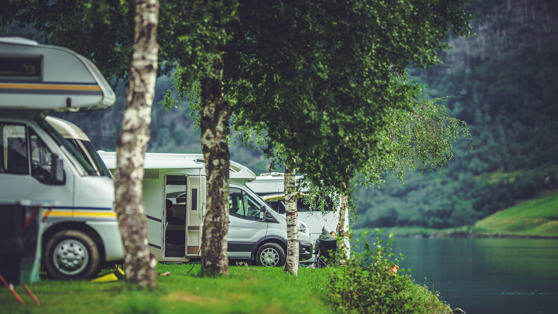 Campground with RVs parked near a lake and trees. Mountains in the background.
