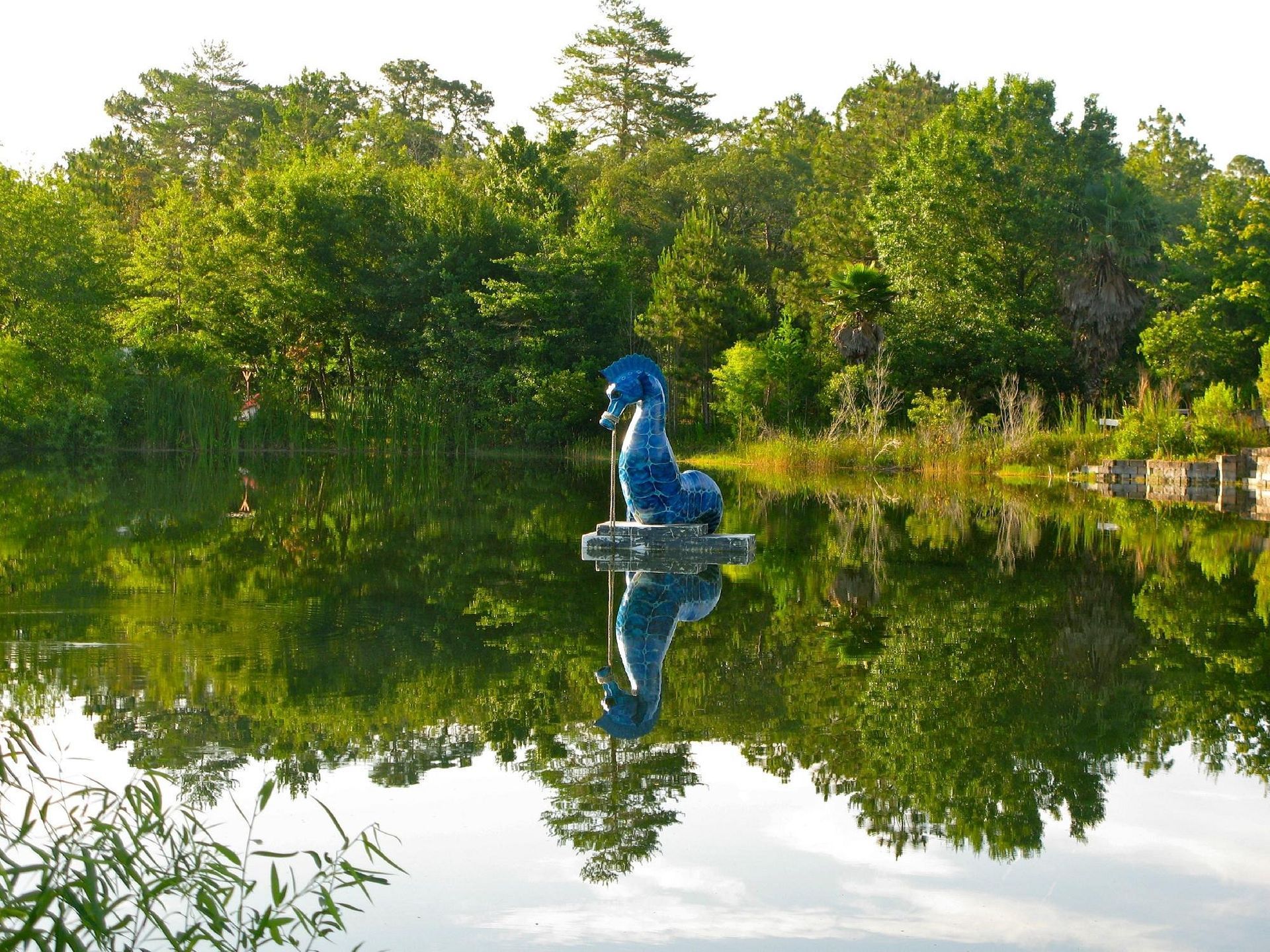 Blue seahorse statue in a lake, reflecting green trees and sky.