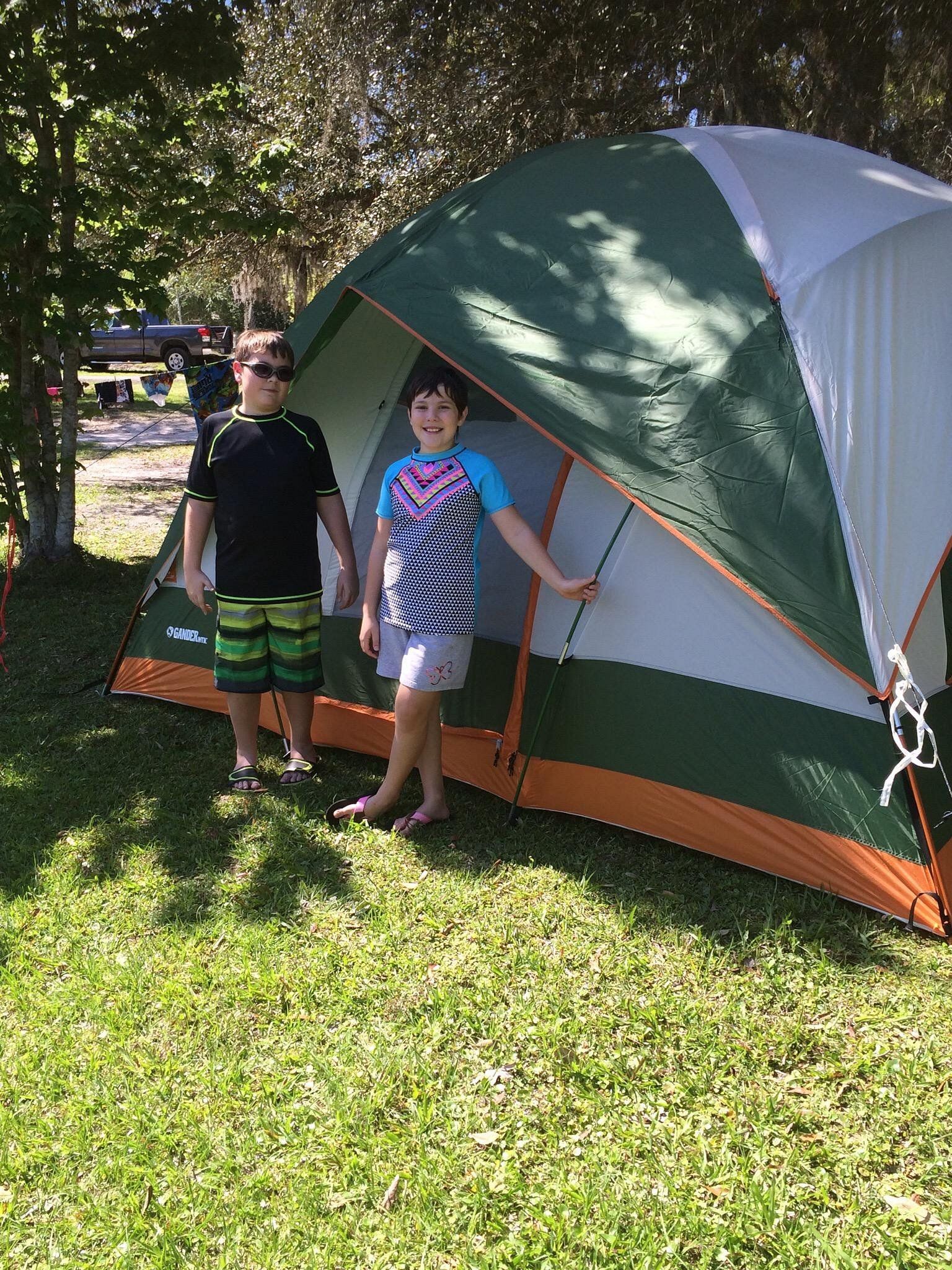 Two children stand outside a green and white tent in a grassy area.