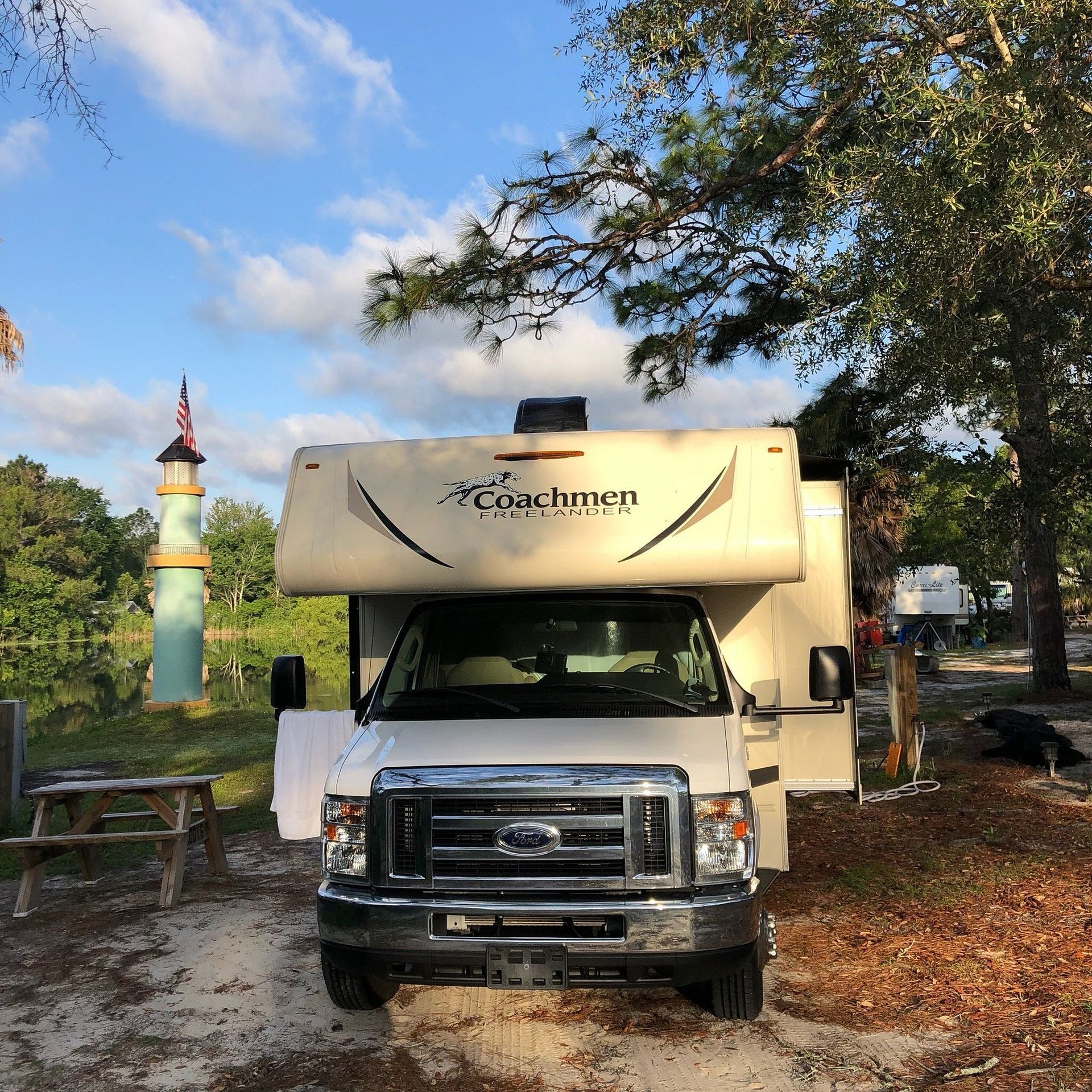 An RV parked at a campsite on a sunny day. A lighthouse stands nearby.