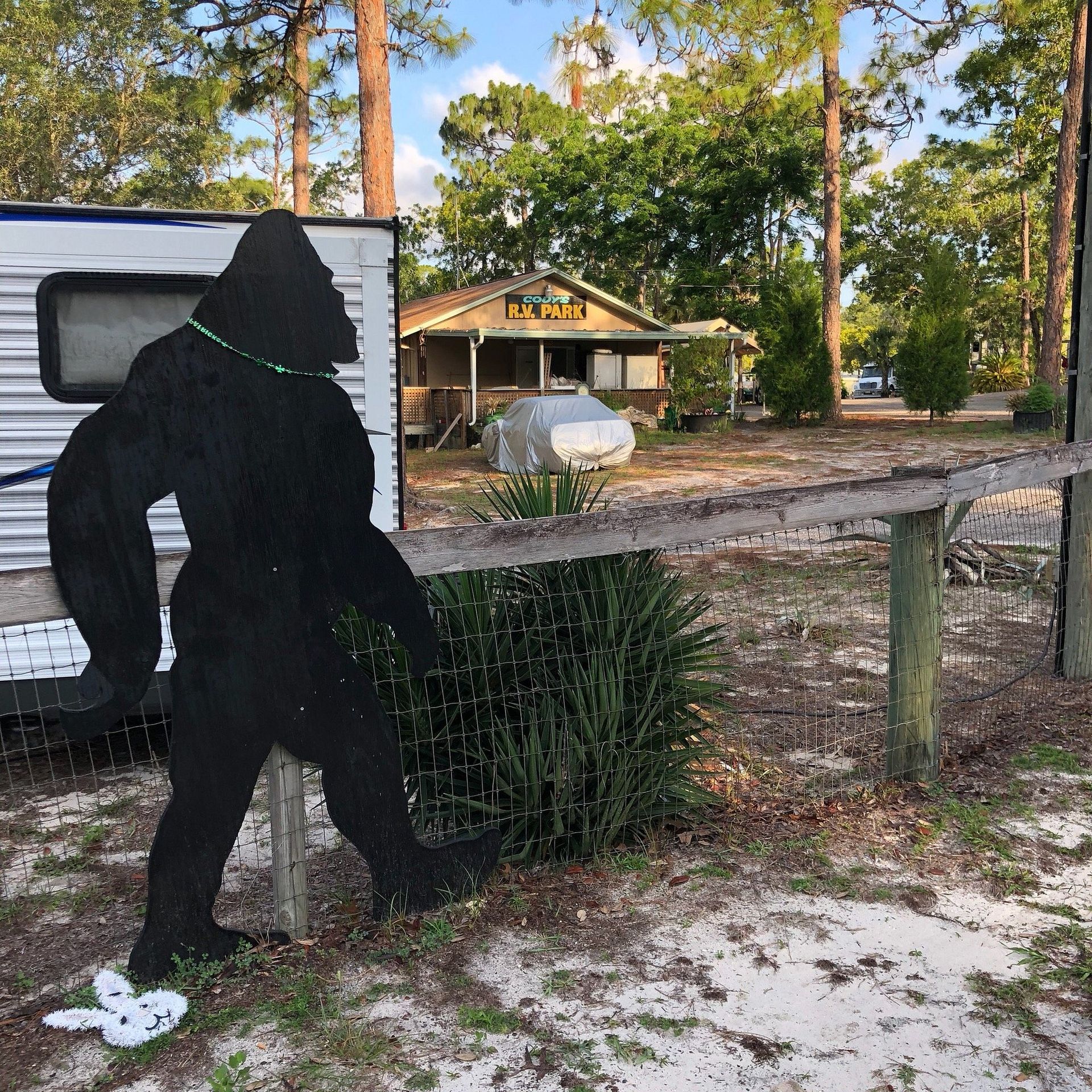 Black silhouette of Bigfoot stands near a fence and trailer; small building with sign in background.