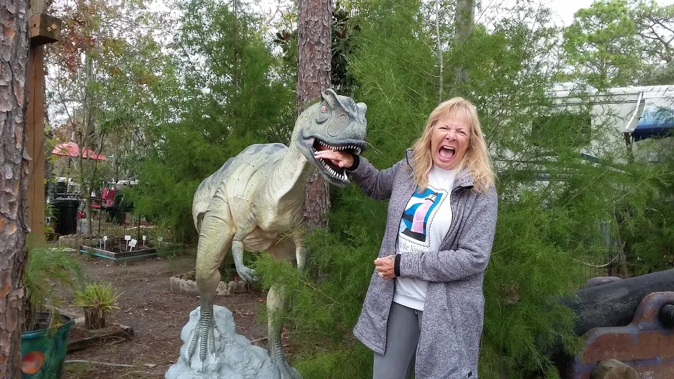 Woman playfully interacts with a dinosaur statue, smiling and laughing outdoors.
