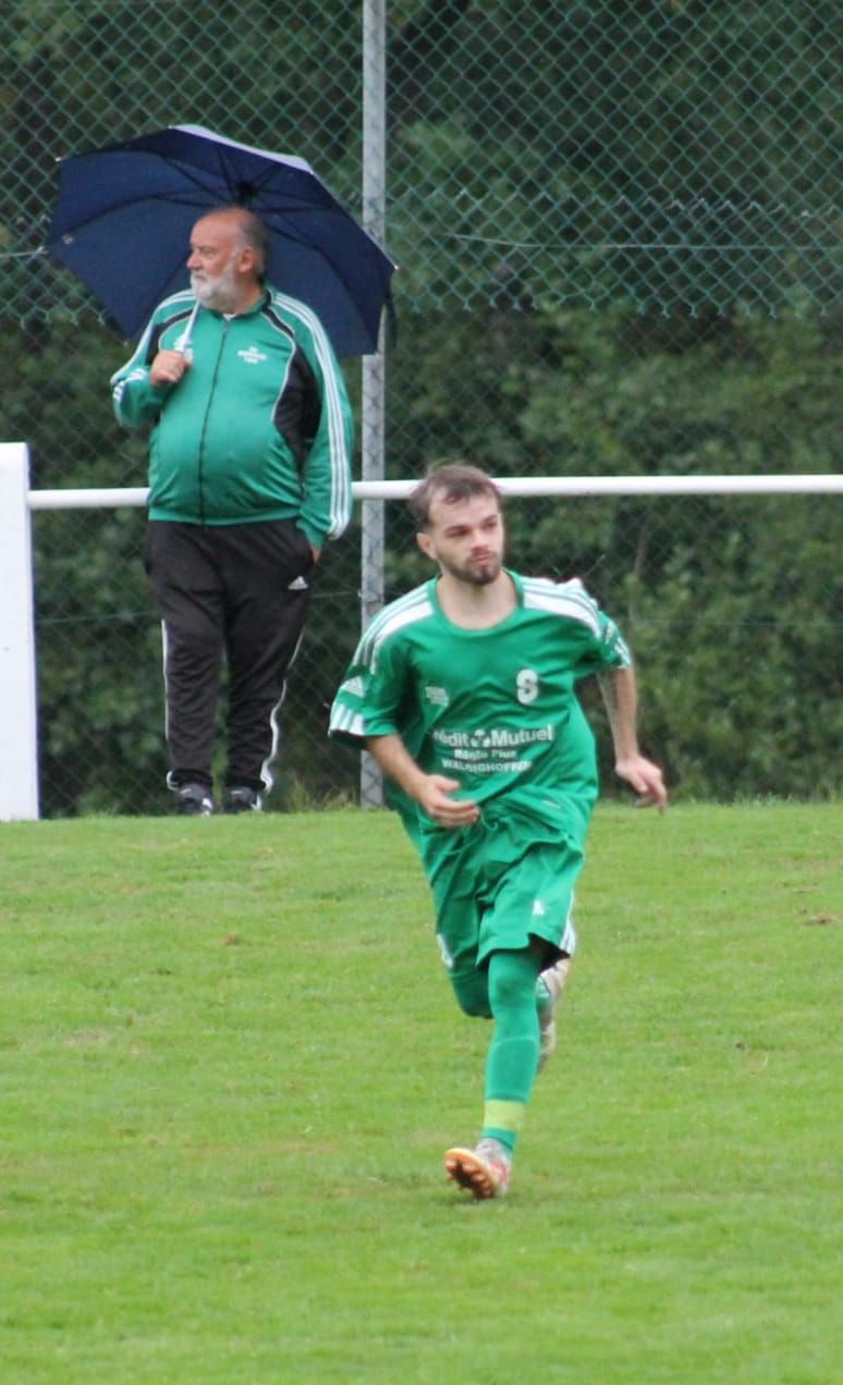 Un homme en chemise verte court sur un terrain de football tandis qu'un autre homme tient un parapluie.