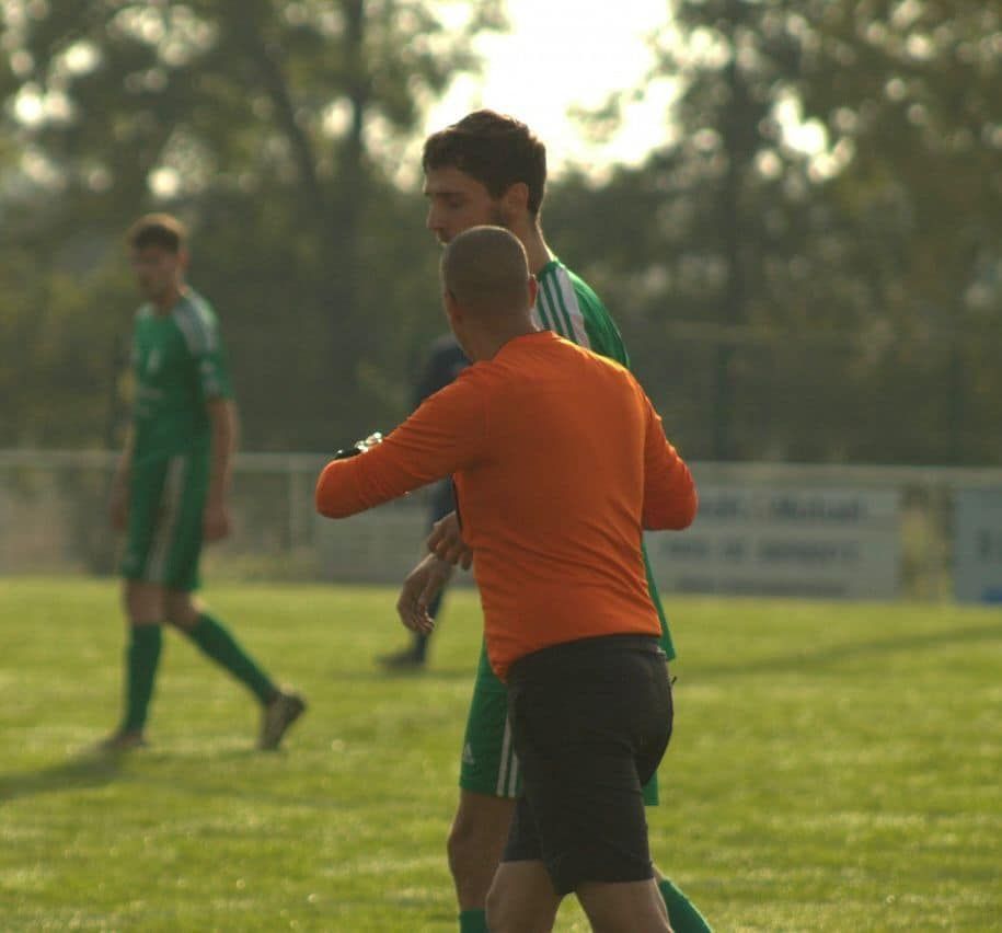 Un homme en chemise orange parle à un homme en chemise verte sur un terrain de football