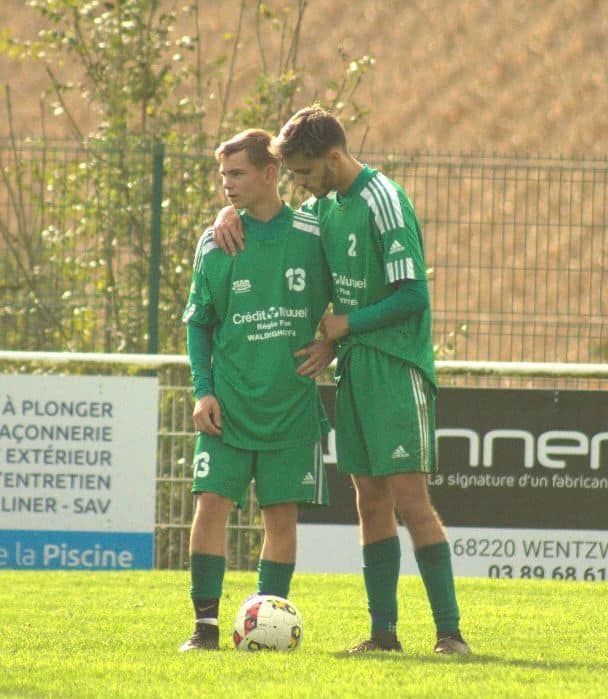 Deux joueurs de football debout sur un terrain avec un panneau en arrière-plan indiquant piscine
