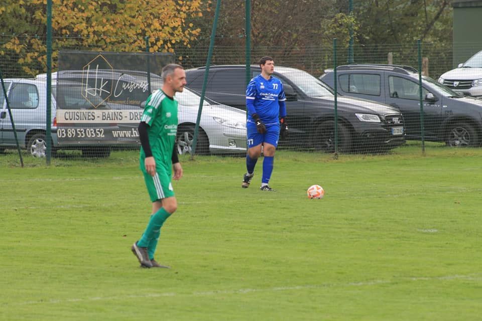 Deux hommes jouent au football sur un terrain avec des voitures garées en arrière-plan.