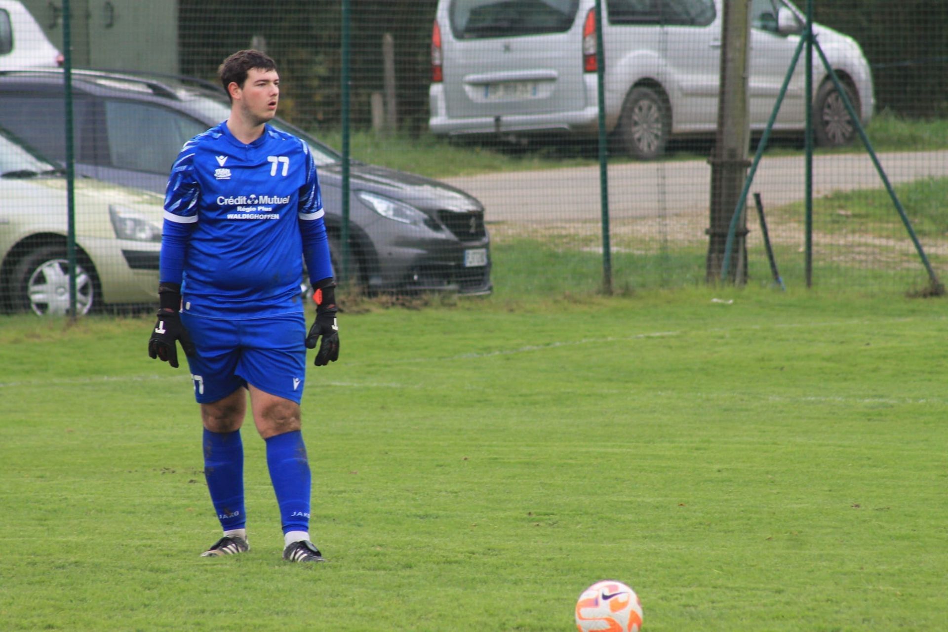 Un homme en uniforme de football bleu se tient à côté d'un ballon de football sur un terrain.