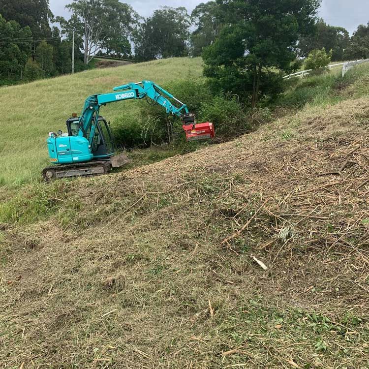 A Blue Excavator Is Cutting Grass On A Hillside — Atholyn Earthmovers in Wyee, NSW