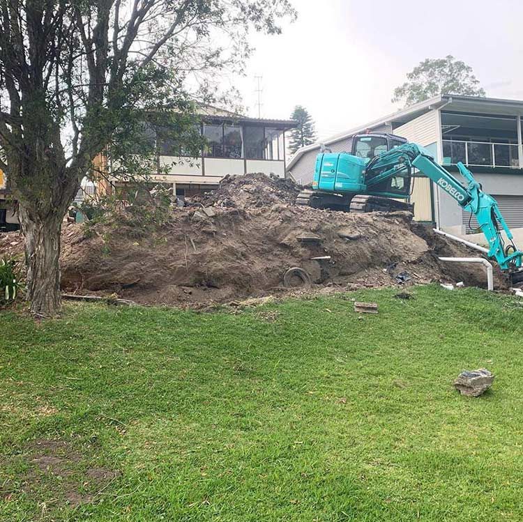 A Blue Excavator Is Sitting On Top Of A Pile Of Dirt In Front Of A House — Atholyn Earthmovers in Wyee, NSW
