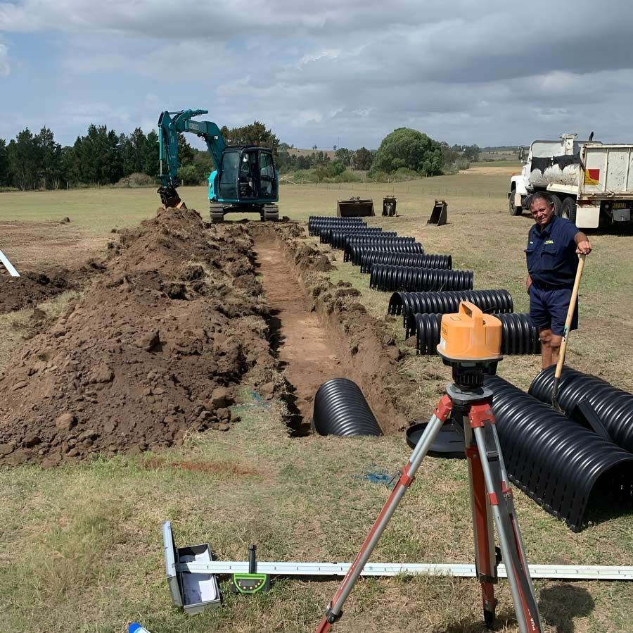 A Man Is Standing In A Field Next To A Row Of Black Pipes And Excavator — Atholyn Earthmovers in Wyee, NSW