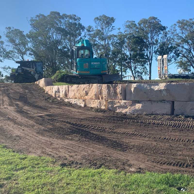 A Bulldozer Is Driving Down A Dirt Road Next To A Stone Wall — Atholyn Earthmovers in Maitland, NSW