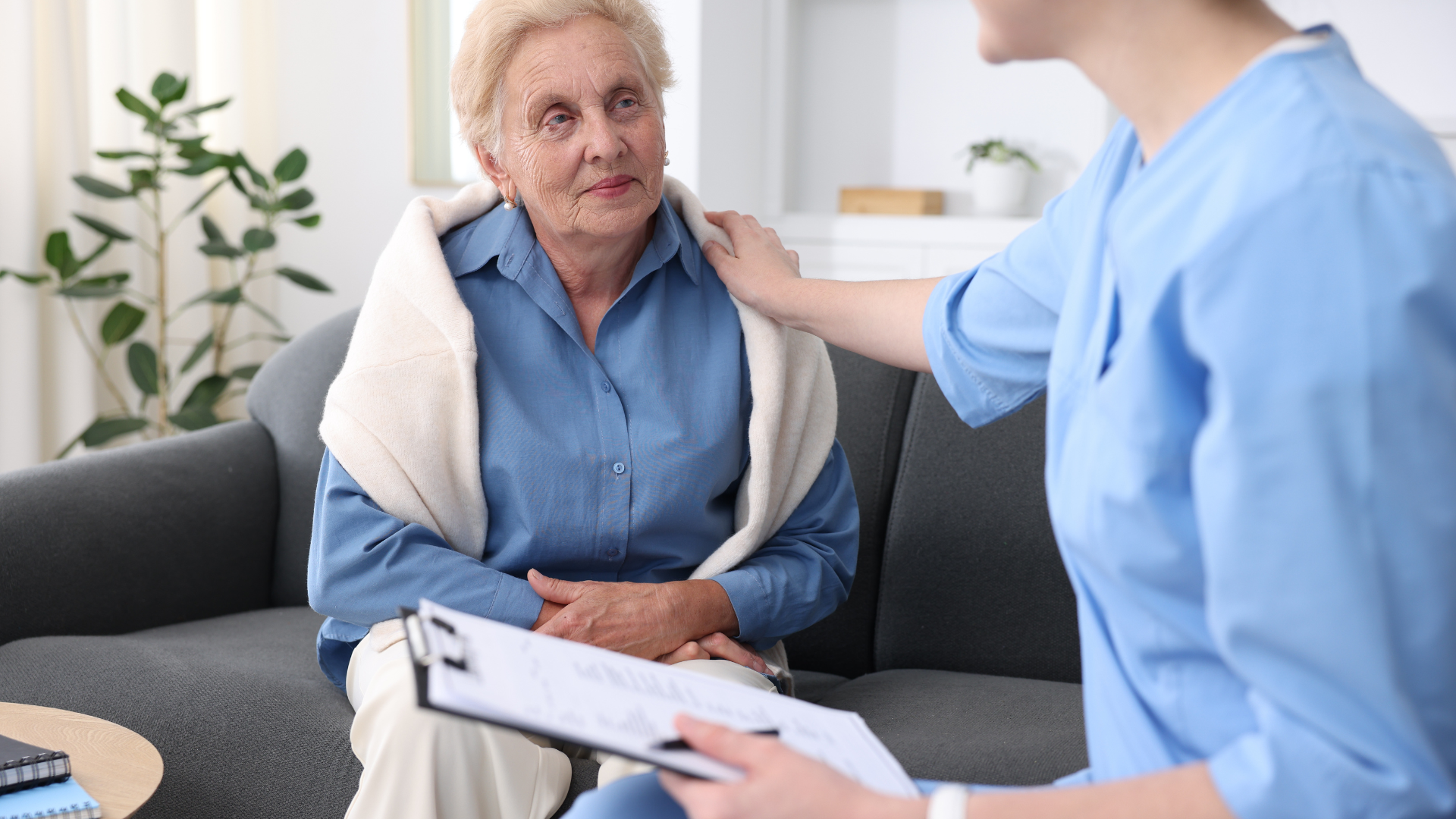 Woman in blue shirt on sofa, comforted by person in scrubs holding clipboard.