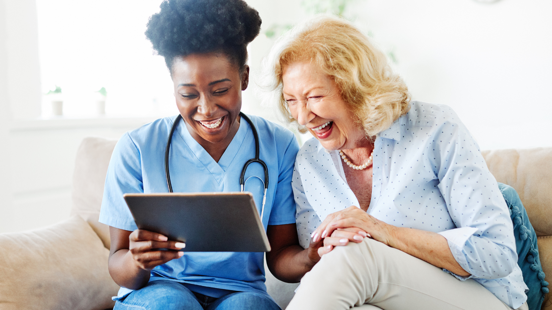 Woman in blue scrubs and elderly woman laughing while looking at a tablet together on a couch.