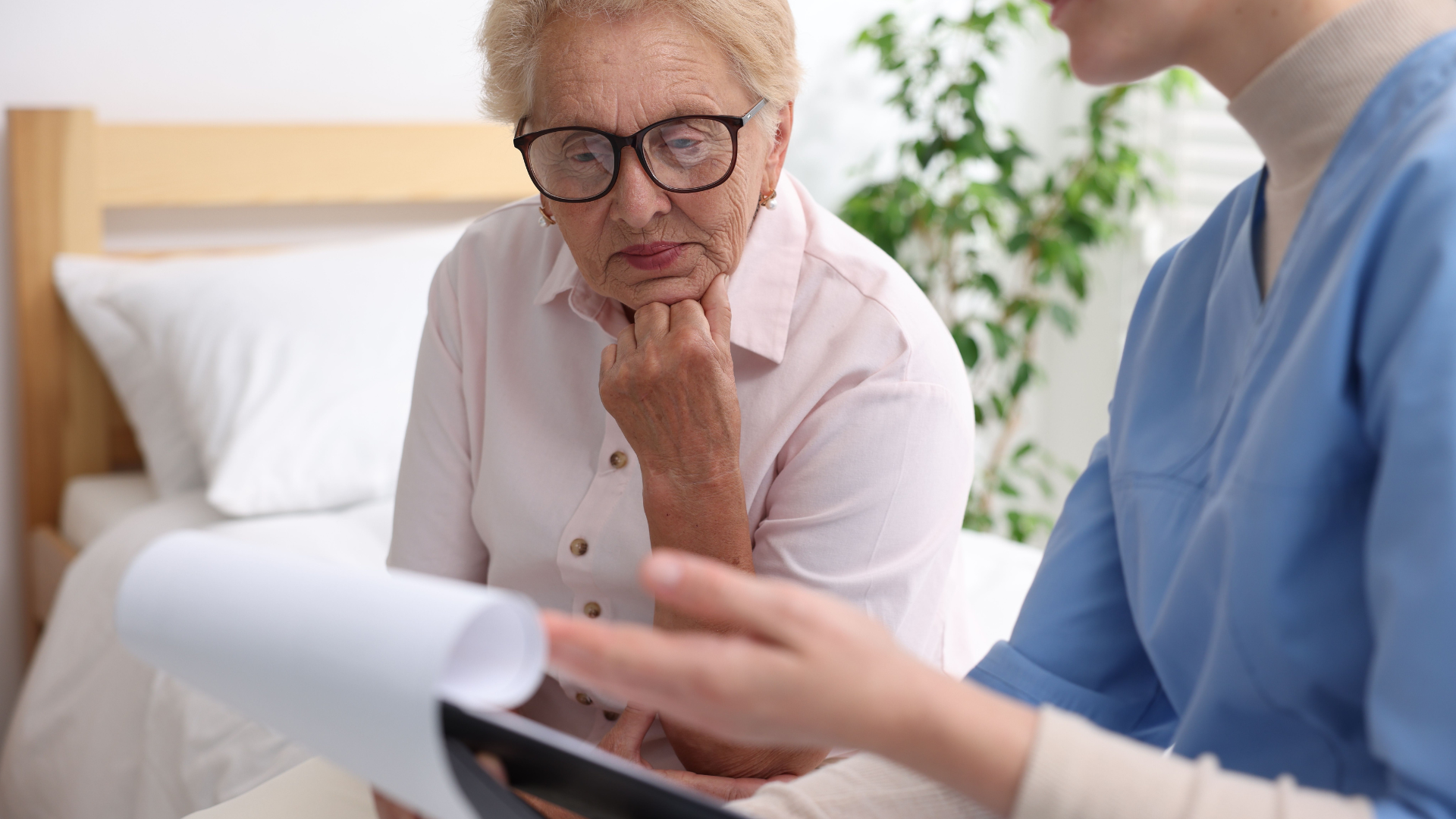 An older adult in glasses listens intently as a healthcare worker in blue scrubs points to a document; bed visible.