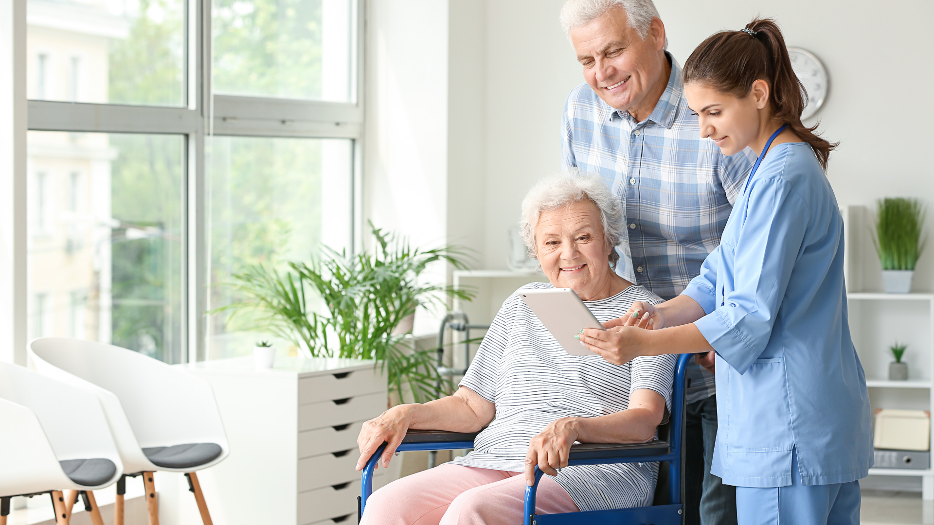 Woman in wheelchair looking at tablet with a caregiver and man; bright room.