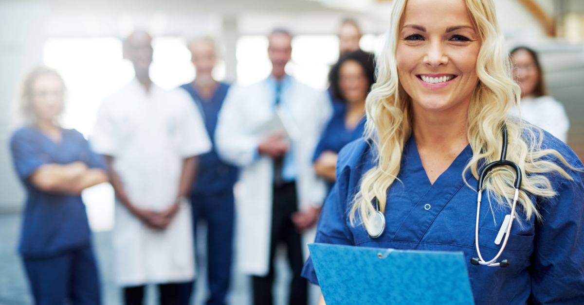 An older adult in glasses listens intently as a healthcare worker in blue scrubs points to a document; bed visible.