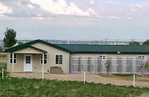 Single-story building with green roof and beige siding, possibly an animal shelter. Set in a grassy field.