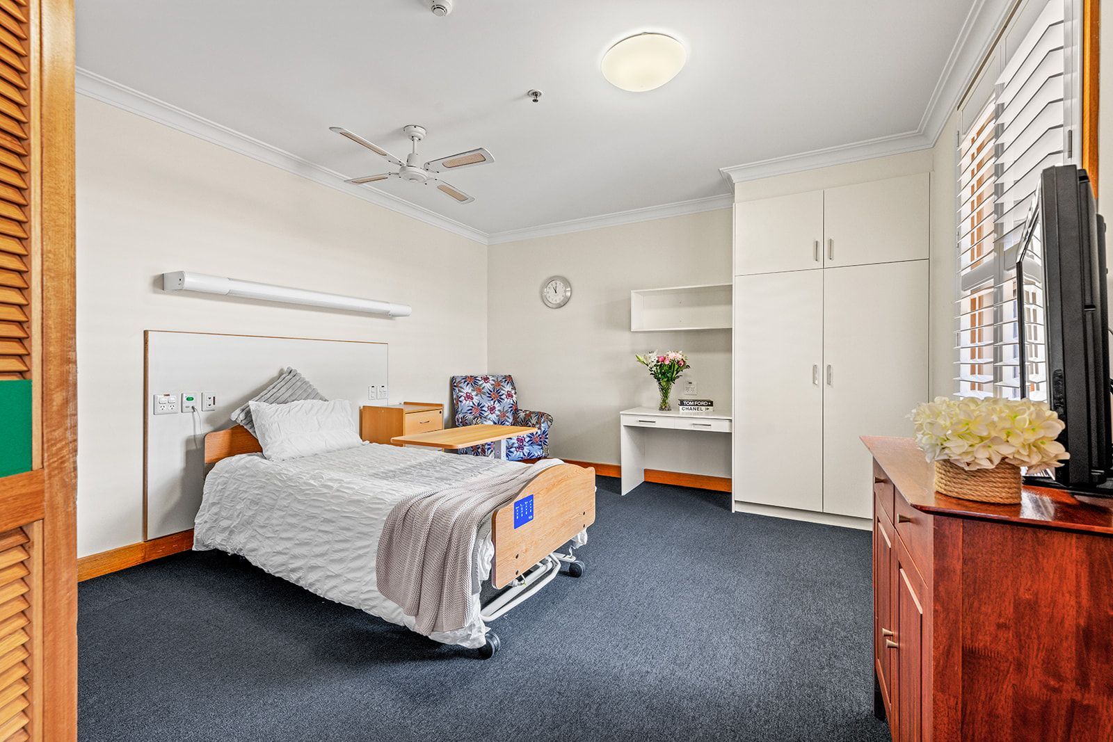 A furnished private room with a single adjustable bed, a desk, a floral armchair, and white wardrobe cabinets.