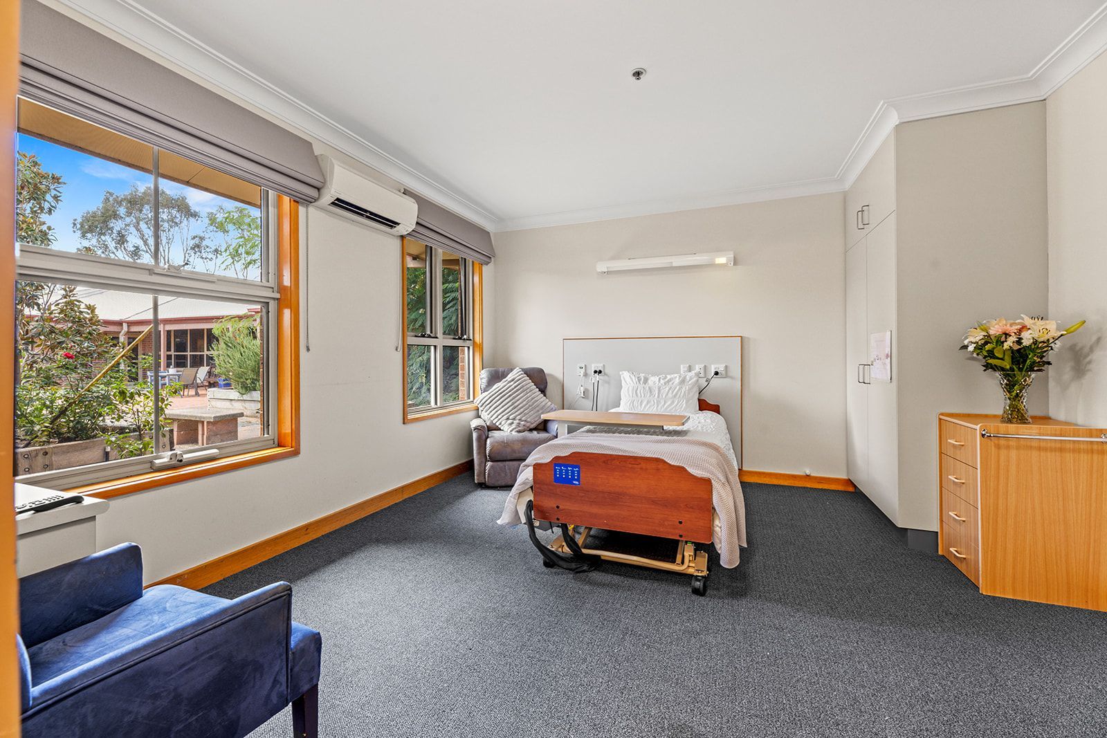 A bright, furnished bedroom in an aged care facility with a hospital-style bed, two armchairs, and a floral arrangement.