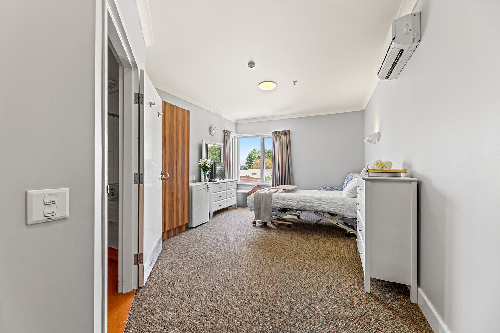 A furnished bedroom with tan carpet, a bed, a white dresser, a closet, and an air conditioning unit on the wall.