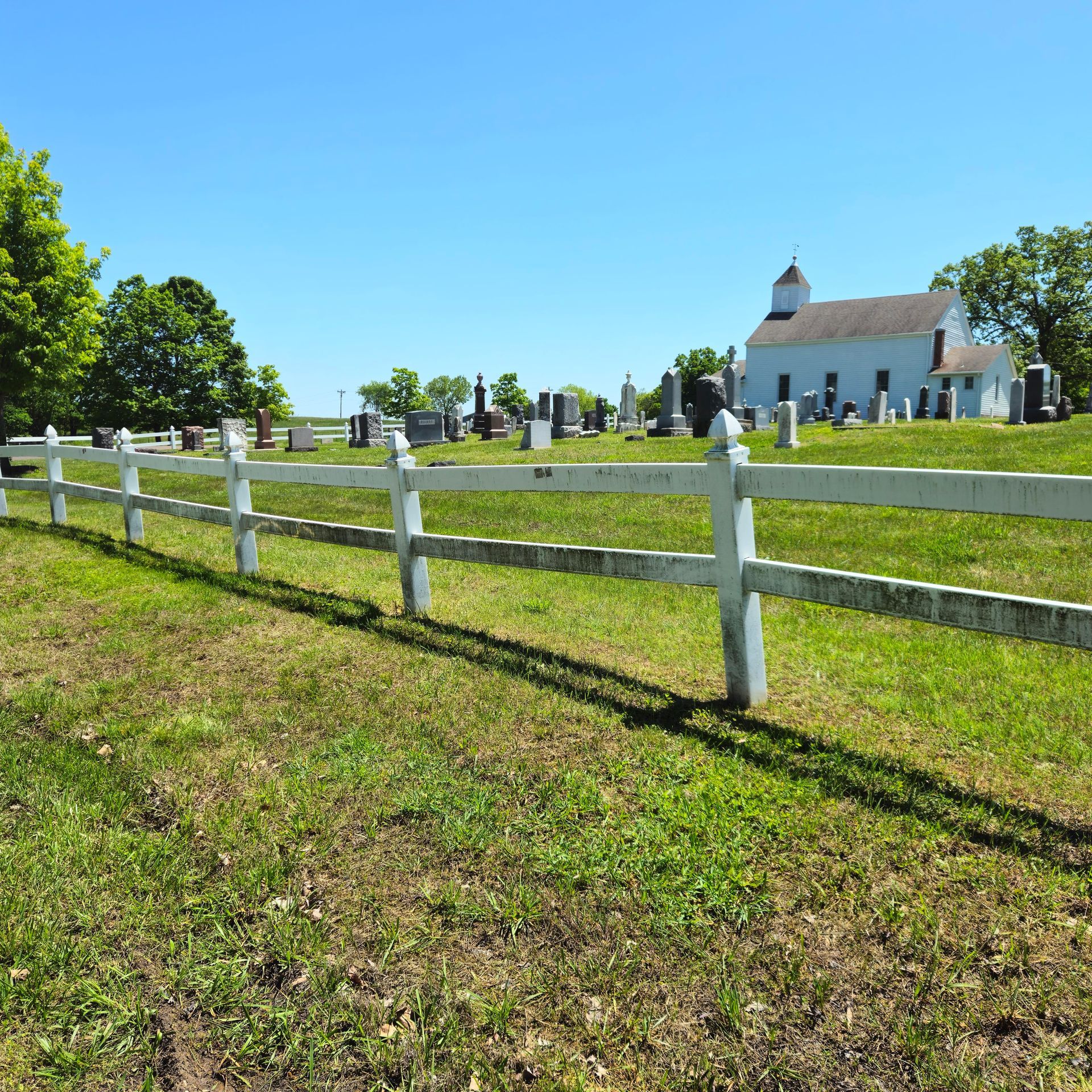 White fence in a green cemetery on a sunny day, with a small church in the background.