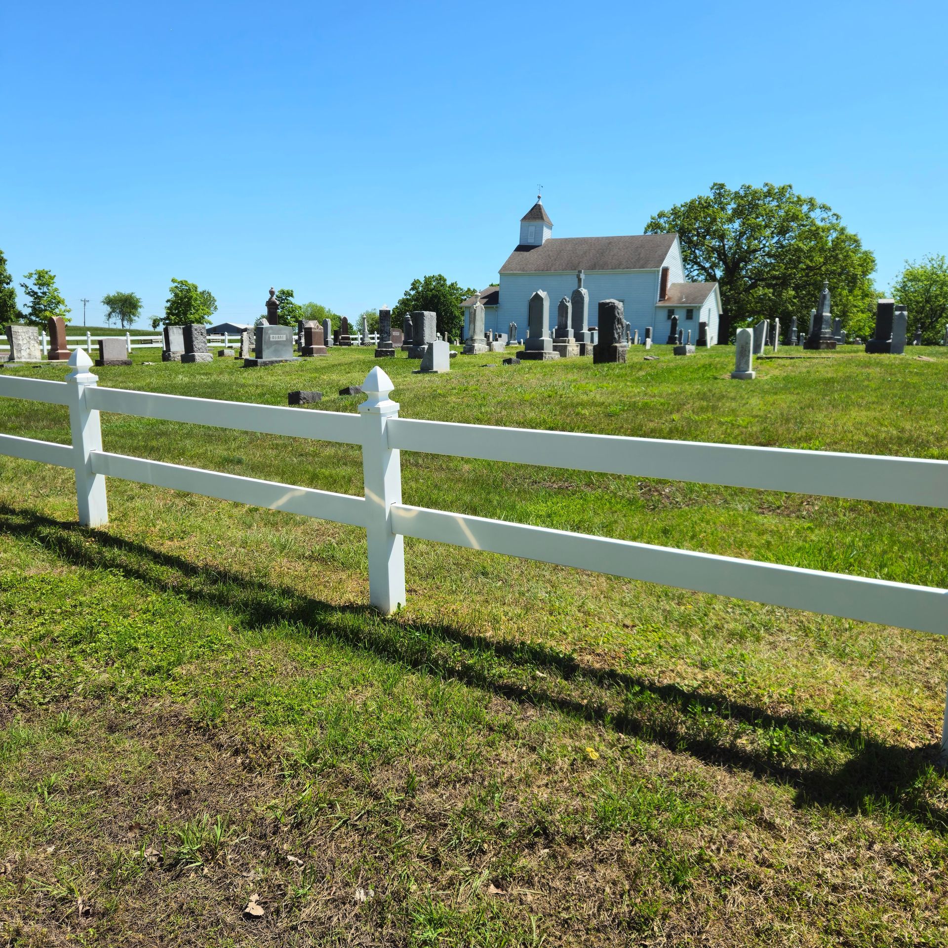 White fence in a sunny cemetery with gravestones and a small chapel in the background.