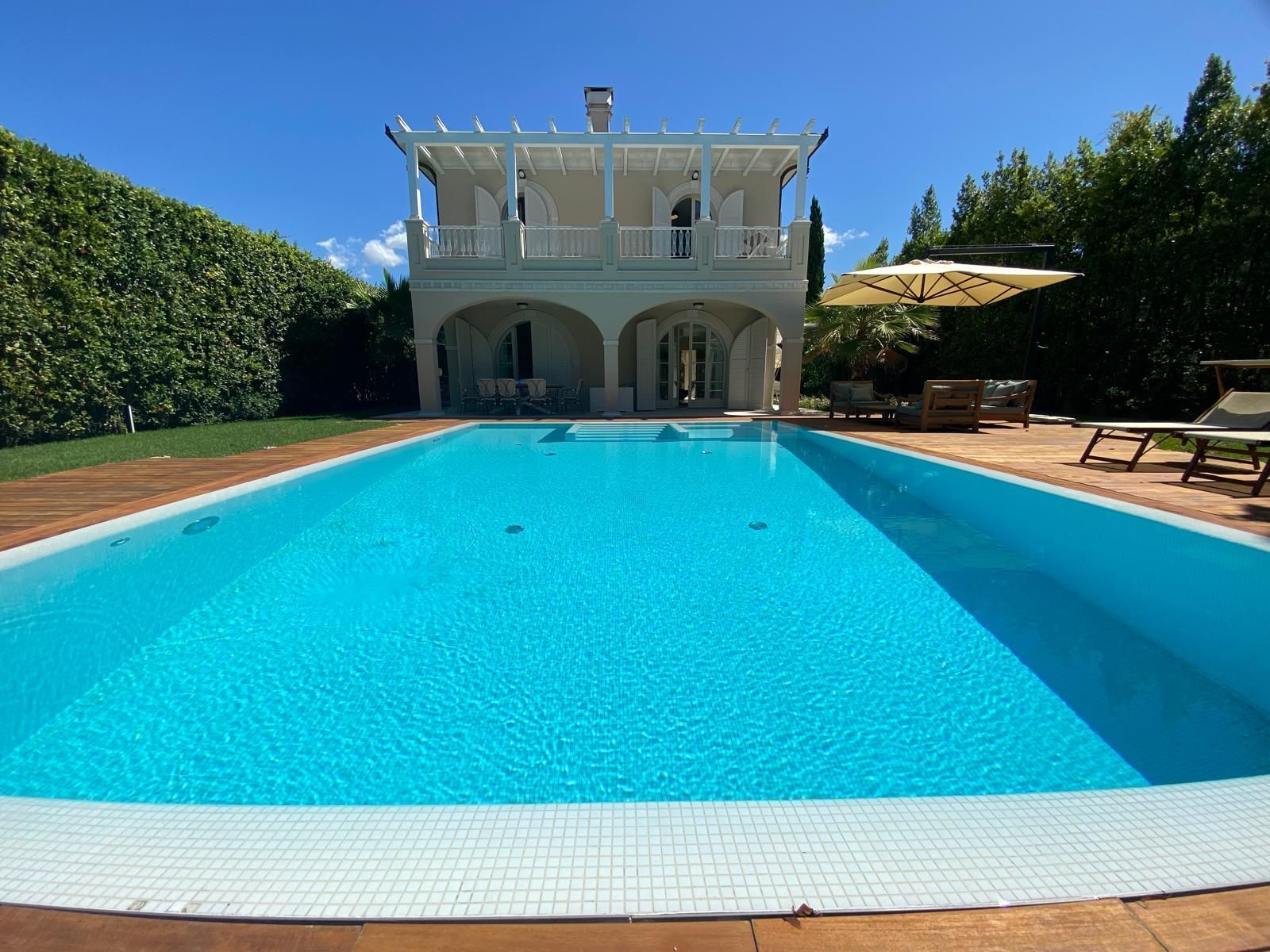 Una piscina turchese brillante di fronte a una villa bianca sotto un cielo azzurro limpido.