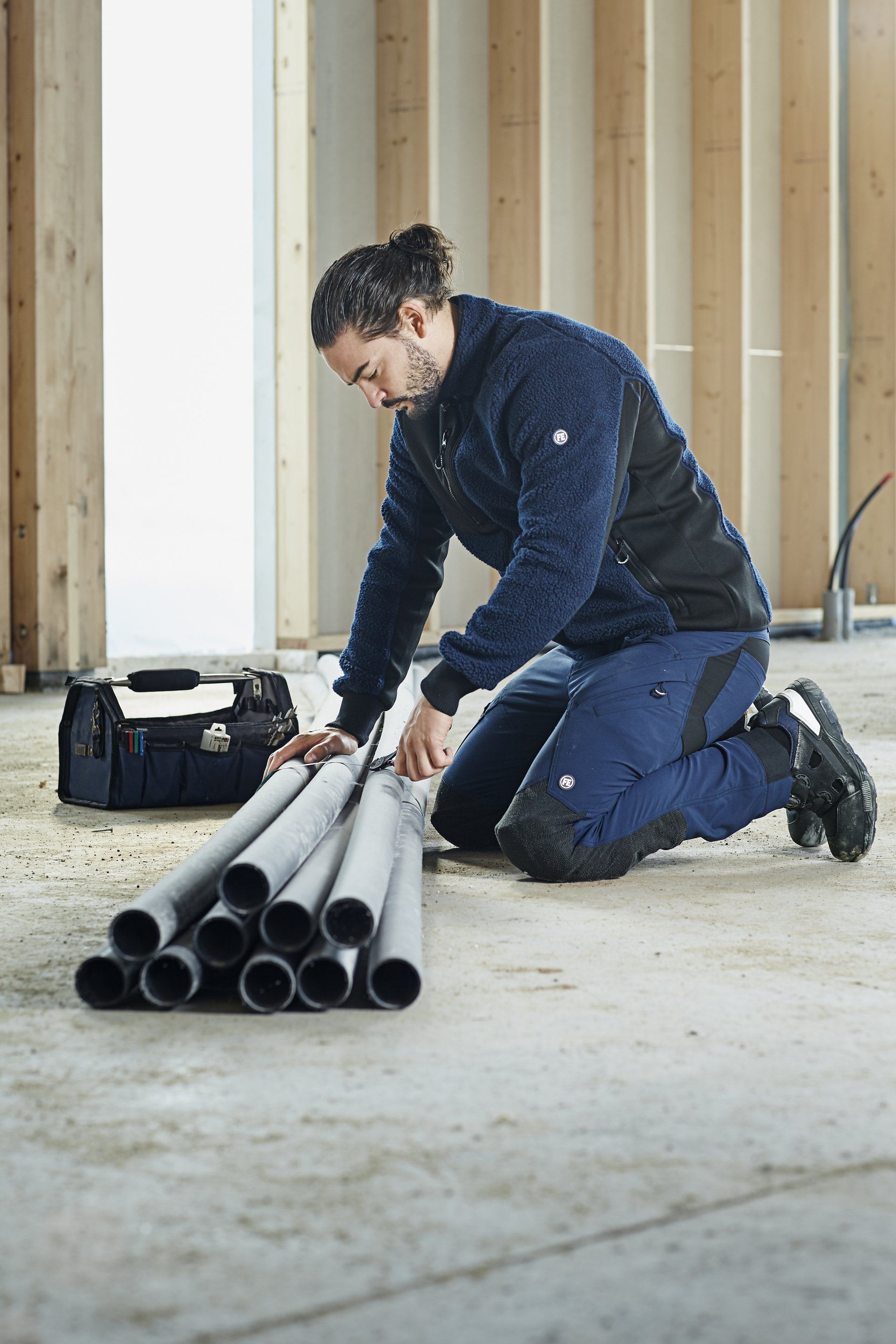 Man kneels in a construction site, examining pipes. He wears a fleece jacket and work pants.