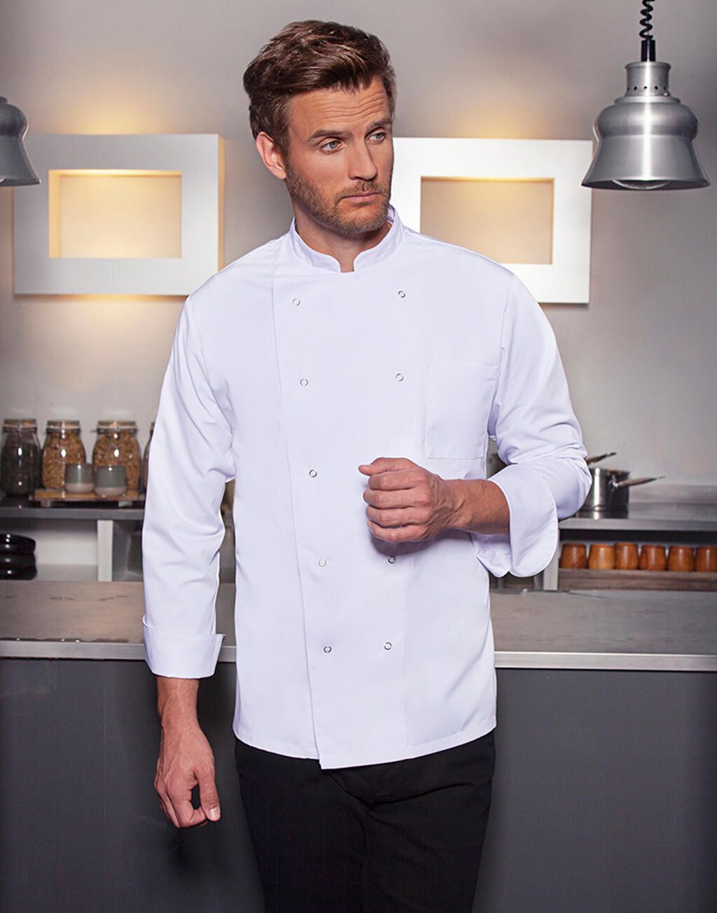 Chef in white coat and black pants in a kitchen, looking to the side.