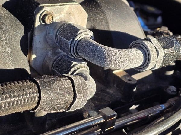 Close-up of black engine hoses and fittings connected to a metal flange with bolts.