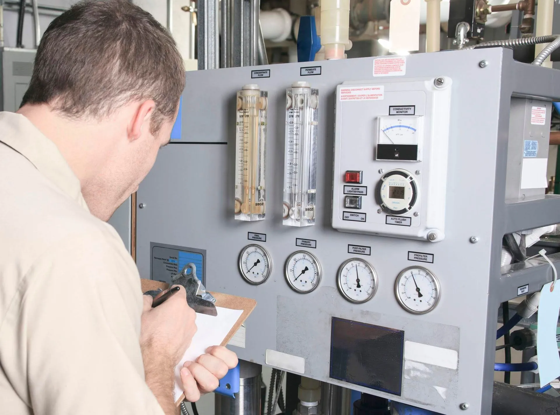 A man is writing on a clipboard in front of a control panel