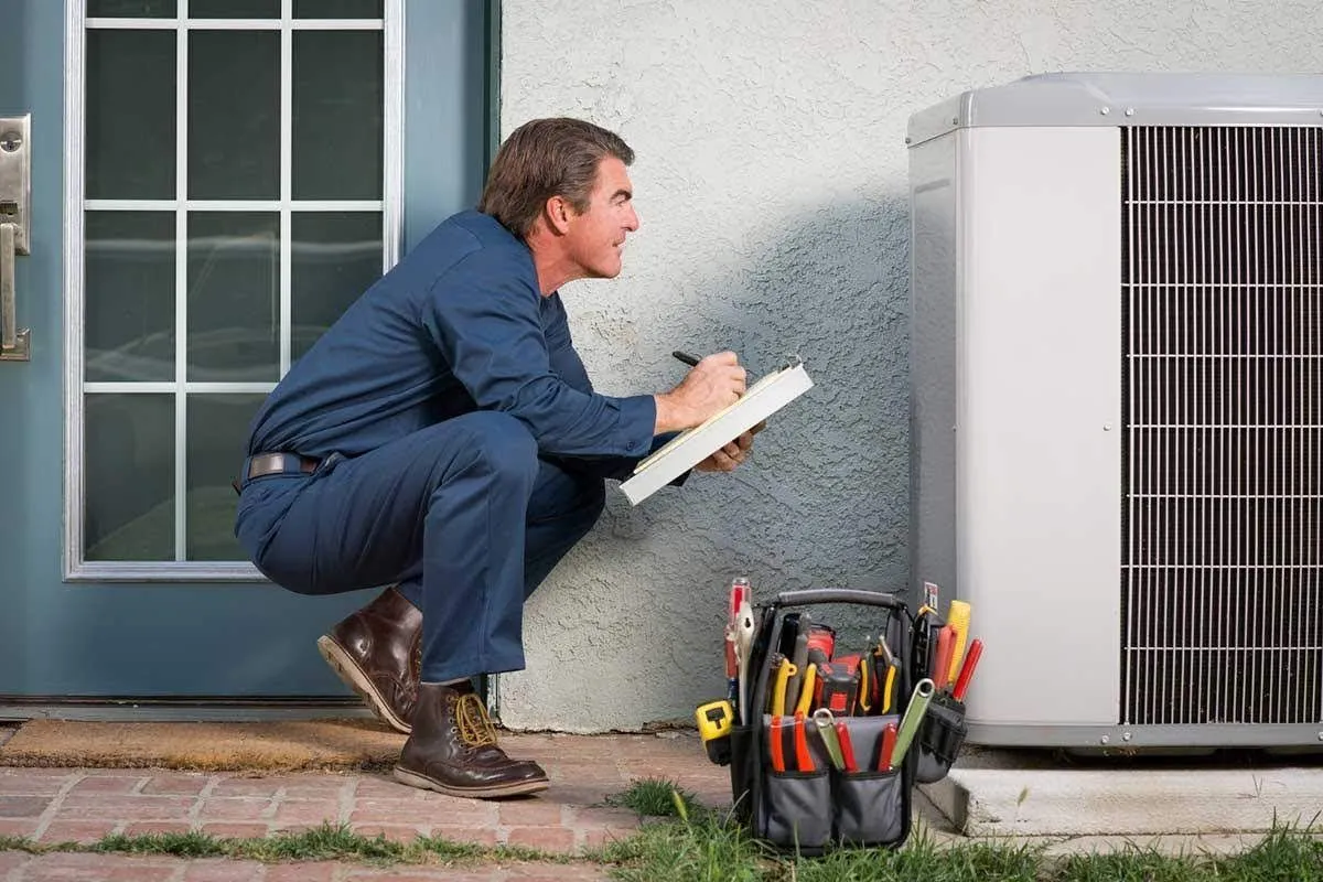 A man is kneeling down next to an air conditioner while holding a clipboard.